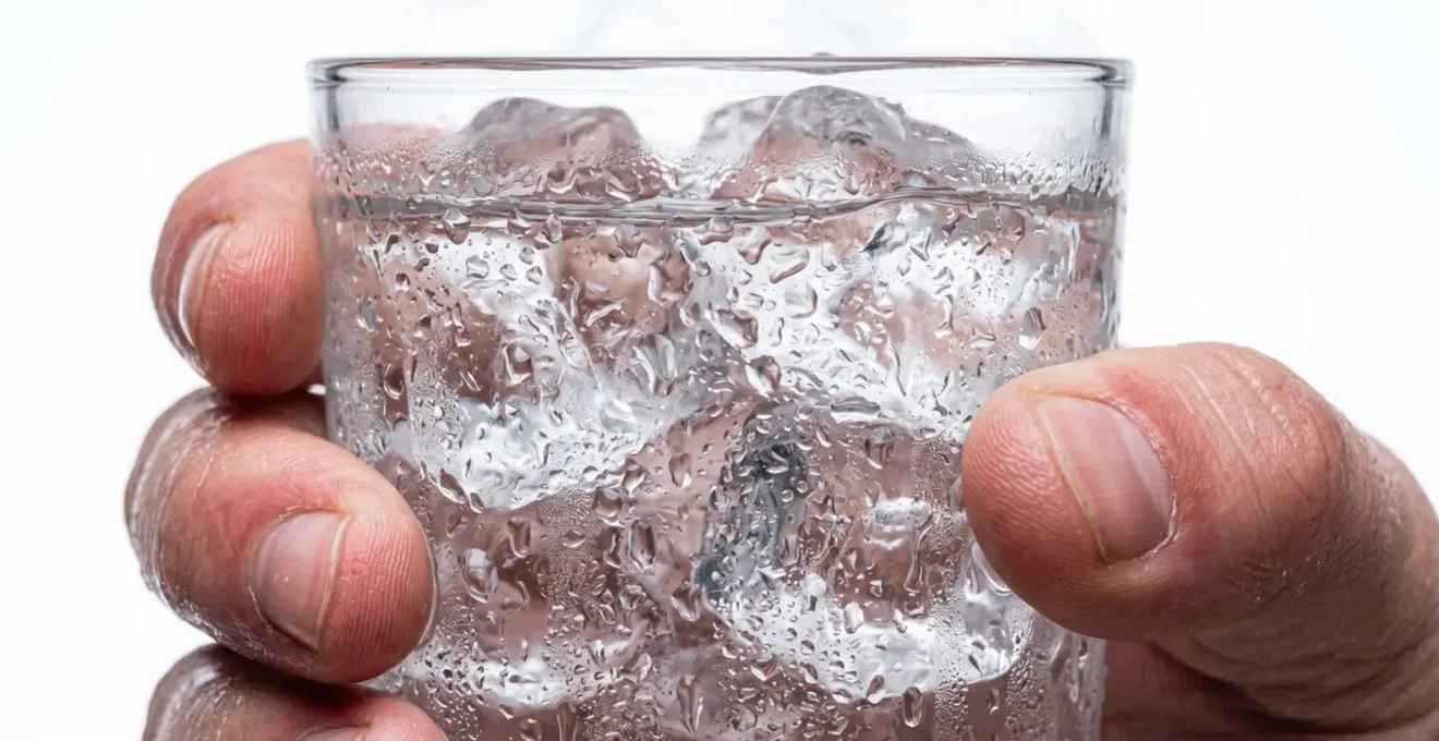 Primer plano de mano sosteniendo vaso cristalino con agua helada y condensación